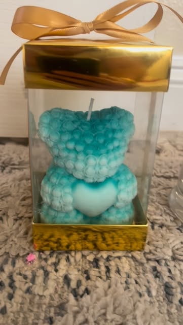 A teal bear candle covered in tiny roses and holding a heart, in a clear gift box with gold ribbon