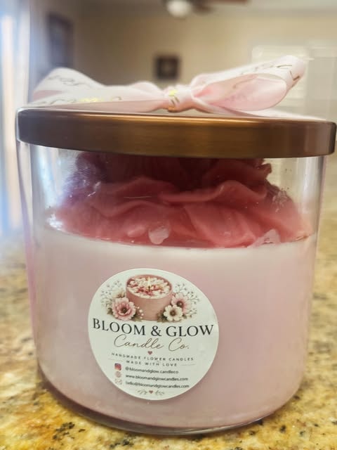 A pink handmade flower candle jar topped with rose petals, a bronze lid and a pink ribbon, with a Bloom and Glow Candle Co. label